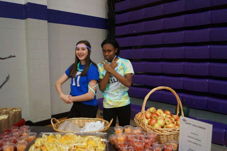 Sophomore Hannah Haworth and sophomore Isley Ford pose in front of the table provided by OPAA Food Management. They handed out oranges, apples, watermelon, and popcorn all for free.